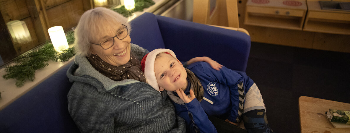 Annemarie Naujeck sitzt mit ihrem Enkel auf dem blauen Sofa. Er zieht eine Grimasse, sie lacht