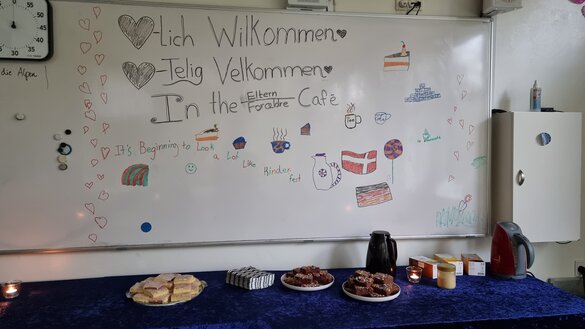 Im Hintergrund ein Whiteboard, mit der Aufschrift Herzlich Willkommen. Sowohl in Dänisch, als auch in Deutsch. Davor drei Kuchen, zwei davon scheinen Brownie zu sein.