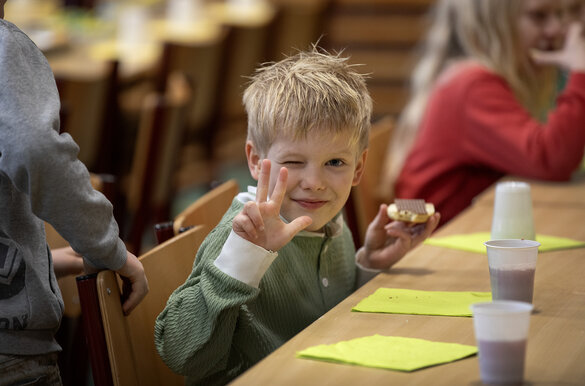 Ein Junge von etwa sieben Jahren steht im Fokus, in der einen Hand sein Brot mit Schokolade, mit der anderen Hand zeigt er die Zahl drei.